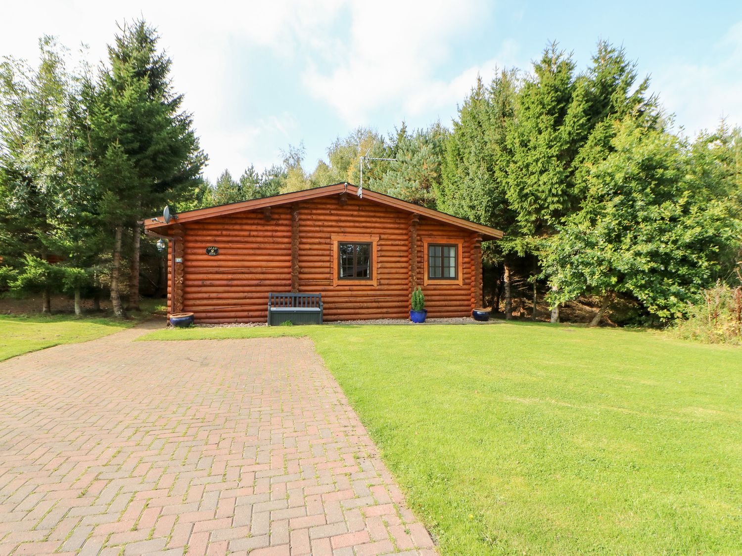 A log cabin with two windows and a bench in front on a paved driveway surrounded by grass and trees at Jalan End in Felton
