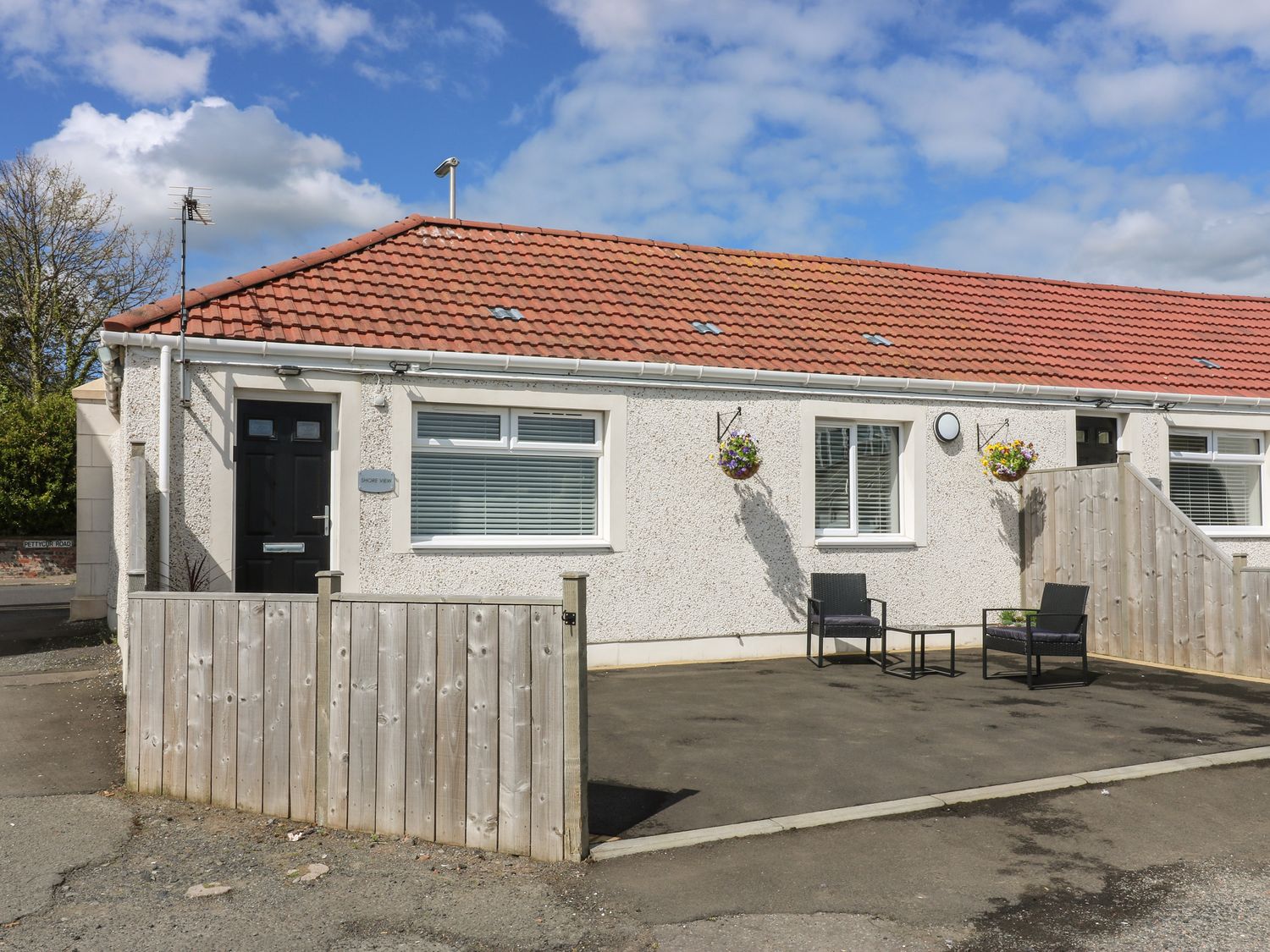 A single story house with a red tiled roof two black chairs and a small table outside and hanging flower baskets at Shore View in Kinghorn