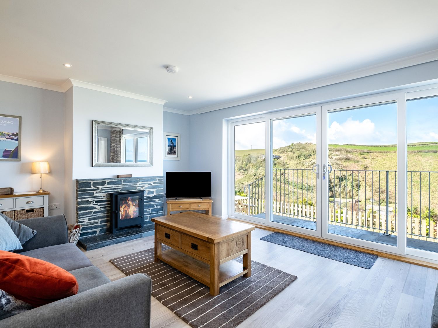 A living room with a sofa a coffee table a fireplace and large glass doors showing a hillside view at Harbour Lights Port Isaac