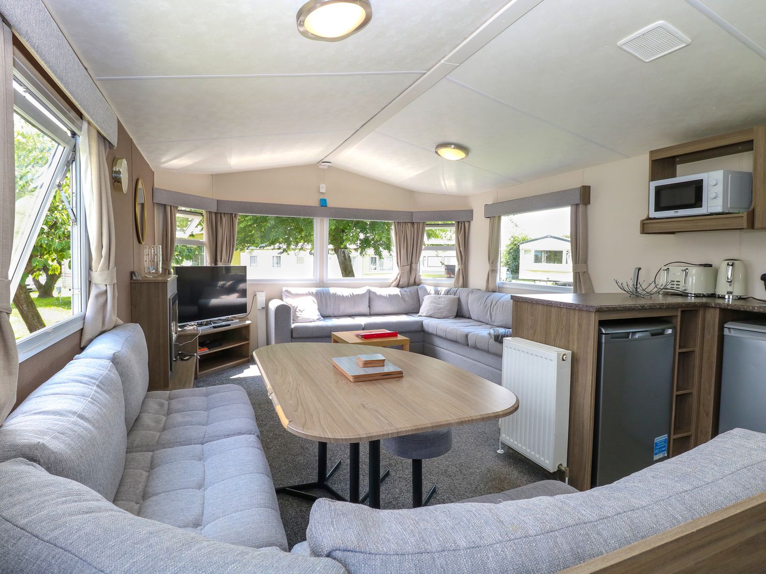 An open-plan living area with grey sofas a wooden table TV and kitchen appliances at 626 in Burgh Castle