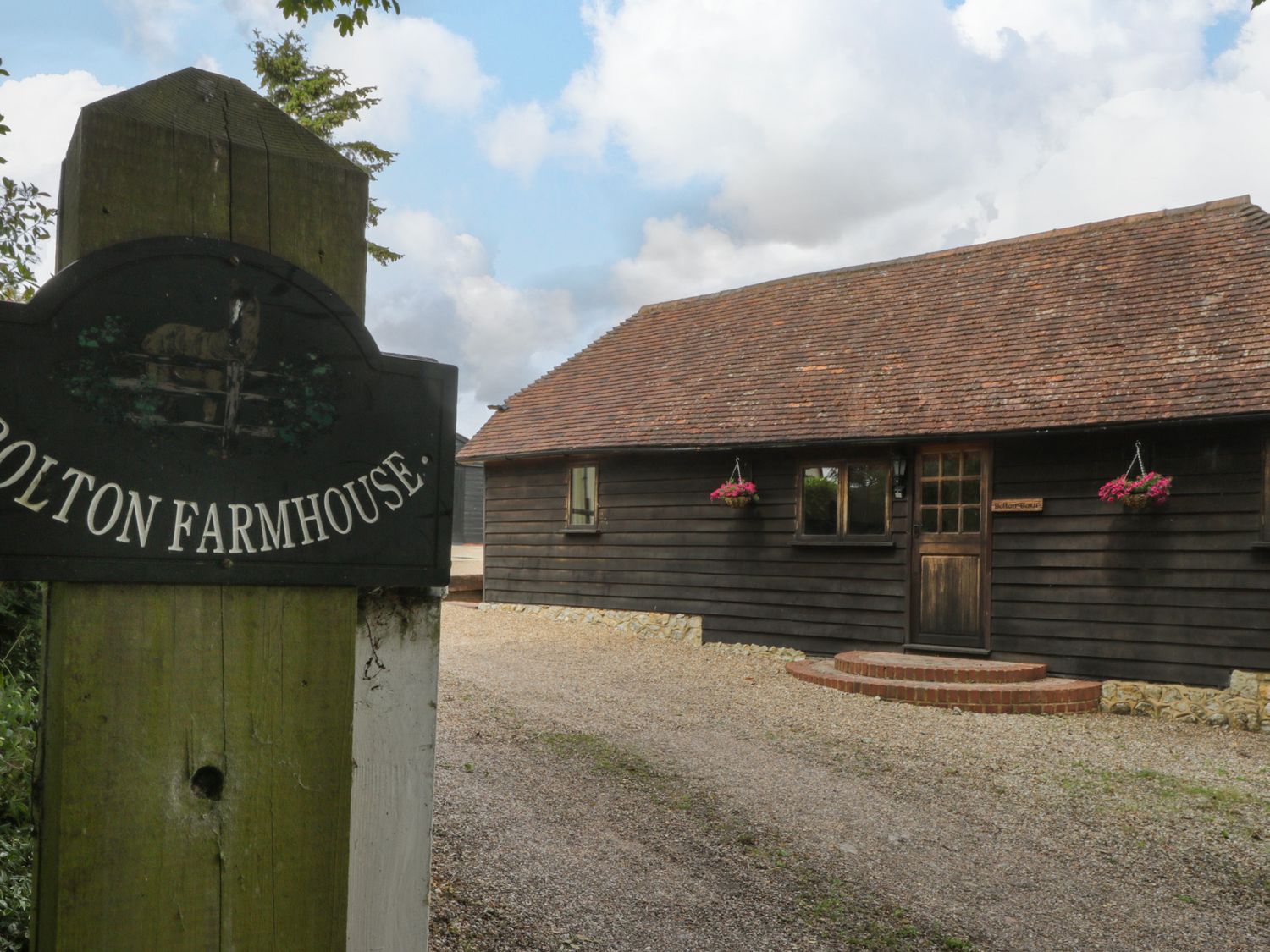 A wooden farmhouse with hanging flower baskets and a gravel driveway with a Bolton Farmhouse sign on a wooden post at Bolton Barn in Lenham Heath near Ashford