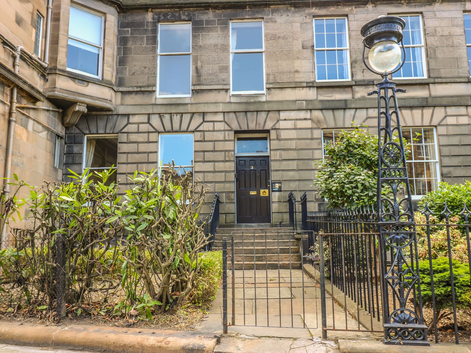 The front entrance of a stone building with steps leading to a black door and a metal gate with surrounding bushes at 3 Lynedoch Place in Edinburgh