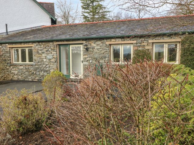 Front view of a stone cottage with three windows and a glass door surrounded by bushes and trees at Apple Tree Cottage in Troutbeck Bridge near Windermere