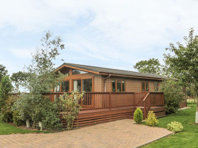 A wooden lodge with a deck and railings surrounded by trees and bushes at Bulmer Farm Lodge in Malton