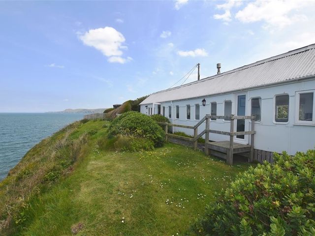 A white single-story building with multiple windows and a small wooden deck beside a grassy area on a cliff overlooking the sea at Clifftop Carriage in Aberporth Ceredigion