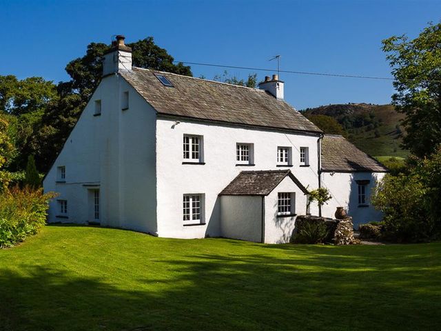 A white two story house with chimneys surrounded by grass and trees at Fair Rigg Old Farm in Cartmel