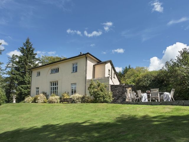 A large house with white walls surrounded by bushes and trees with an outdoor table and chairs on a stone patio at Tent Lodge in Coniston