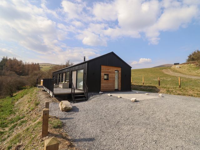 A black and wooden rectangular building on a gravel area with a deck and railing in a rural setting at The Sheep Shed in Llanrhaeadr-Ym-Mochnant