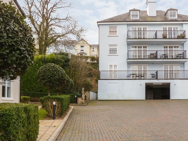 A paved driveway leading to a light blue apartment building with balconies and outdoor furniture at 12 Combehaven in Salcombe