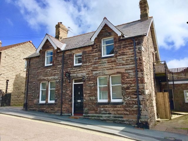 A stone house with white framed windows and a black door on a sloping street at Cheviot Hills in Wooler