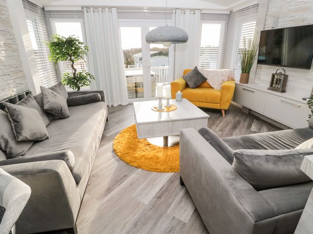 A living room with grey sofas a yellow armchair a white coffee table and a wall-mounted TV at 23 Valley View in Swardland near Felton