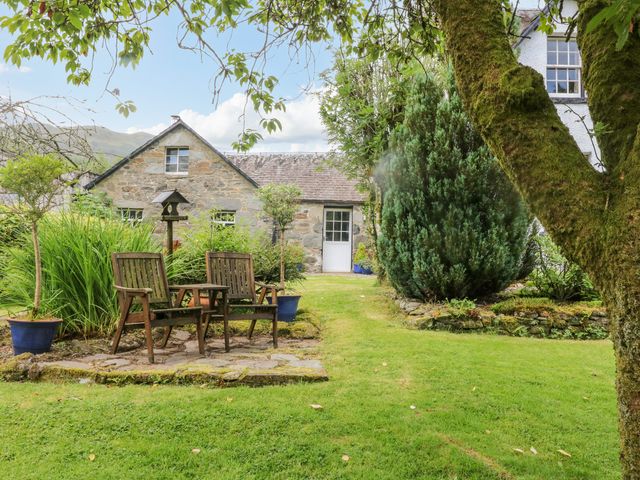 A garden with wooden chairs and a table near a stone cottage surrounded by trees and plants at Inglis Nook Cottage in Balquhidder Perthshire Scotland