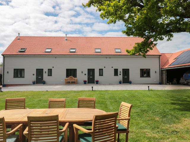 A wooden outdoor dining table with chairs on a lawn in front of a large building with red tiled roof at The Stables in Watchfield near Highbridge