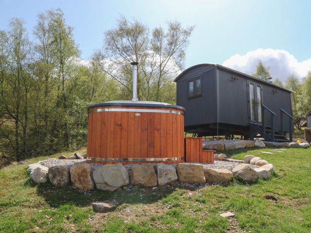 A wooden hot tub surrounded by stones next to a black cabin in a grassy area with trees in the background at Coraldie in Laggan near Newtonmore