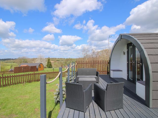 A wooden deck with outdoor wicker furniture next to a curved wooden pod in a fenced grassy area at Cherry Pod 2 Hookagate near Meole Brace