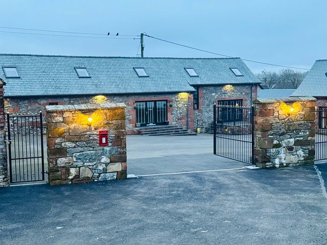 A stone building with a slate roof behind open metal gates and a stone wall with lights and a red post box at The Steading in Terregles near Dumfries