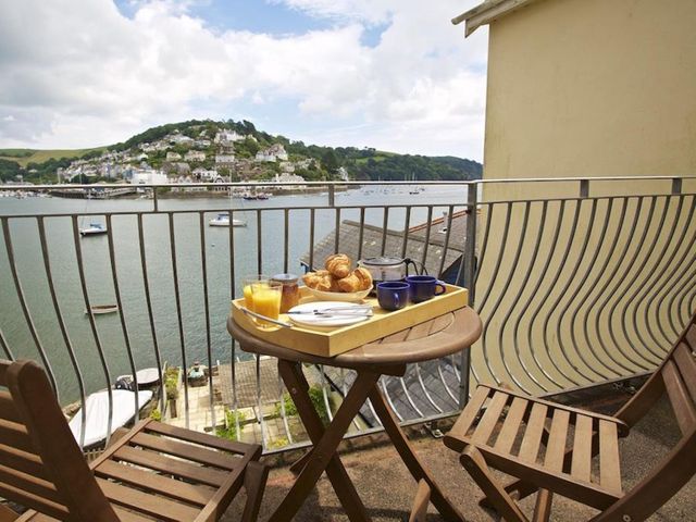 A small balcony with two wooden chairs and a round table holding a tray of breakfast items overlooking a river with boats and houses on a hill at Harbourside in Dartmouth