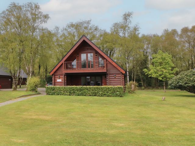 A wooden cabin house with a balcony surrounded by trees and a large grassy lawn at Holly in Dolton