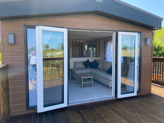 A wooden deck with open glass doors leading to a living area with a sectional sofa and coffee table at Magnolia Lodge Whitby