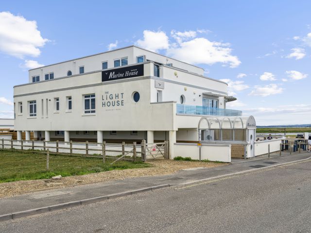 A white building with signs reading Marine House apartments and The Light House seaside bar dining by a road and fenced grassy area at Blue Horizon in Milford On Sea