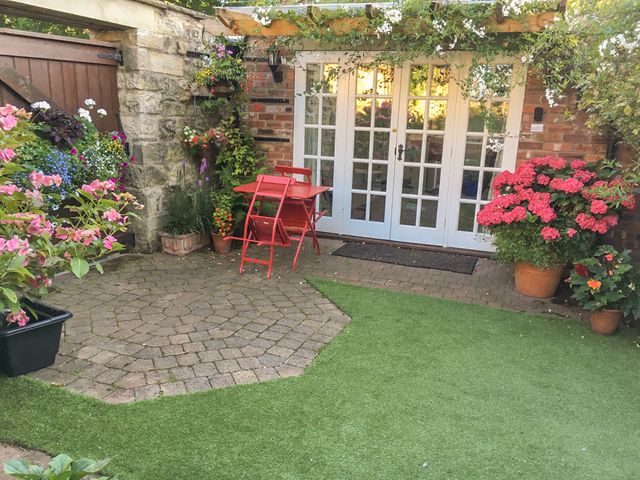 A patio with a red table and chairs surrounded by potted flowers and green artificial grass in front of white French doors at Beech View in Malton