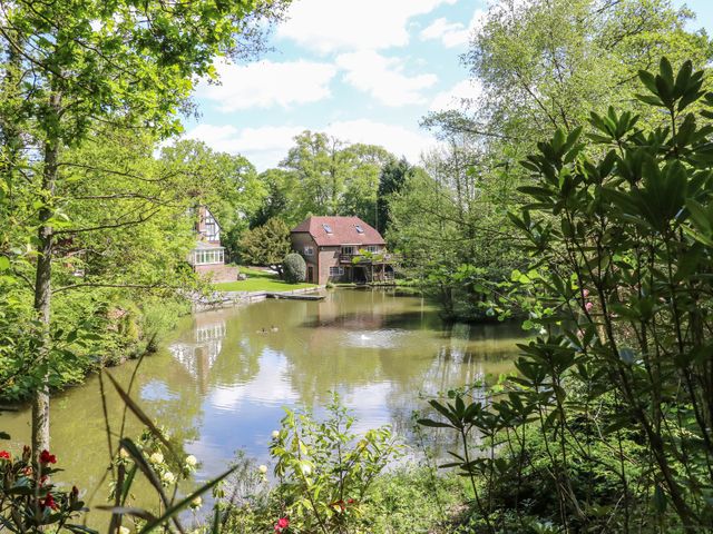 A pond surrounded by trees and bushes with houses on the far side at Miswells Cottages - Lake View in Turners Hill near East Grinstead