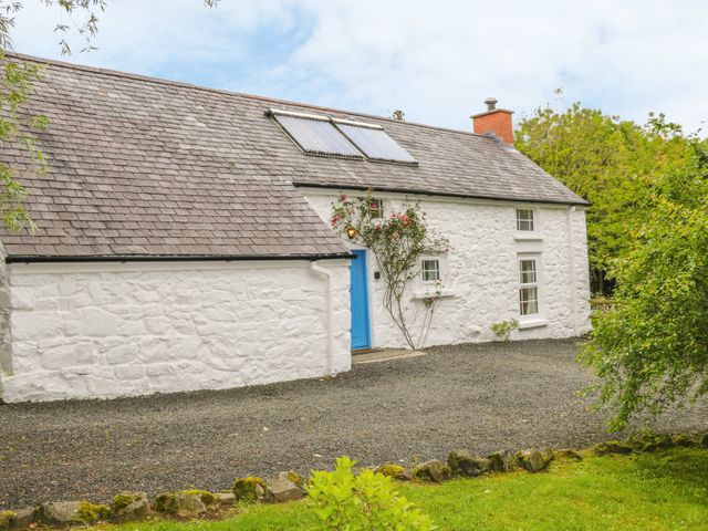 An exterior view of a white stone cottage with a blue door solar panels on the roof and surrounded by greenery at Rosslare Cottage in Broughshane