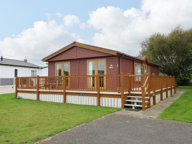 A brown single-story cabin with wooden deck and railing on a grassy plot at Jolie in Hunstanton