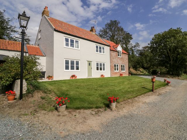 A house with white and brick walls and a tiled roof surrounded by a lawn with potted red flowers at West Farm in Caister-On-Sea