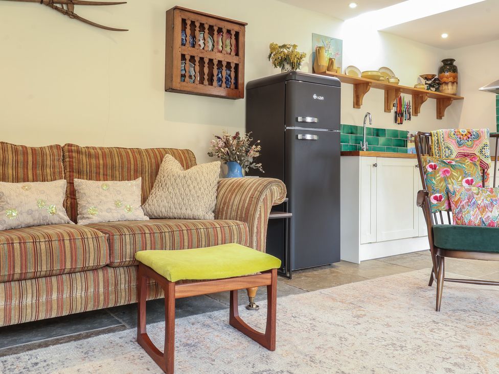 A living area with a striped sofa beside a green cushioned stool and a dark fridge near a kitchen counter at Courtyard Cottage Poplar Farm Barn Sweffling near Saxmundham