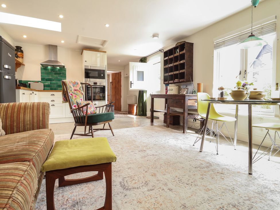 A living space with a striped sofa green cushioned stool wooden chair kitchen cabinets oven green tiled backsplash table with yellow chairs and window light at Courtyard Cottage Poplar Farm Barn Sweffling near Saxmundham