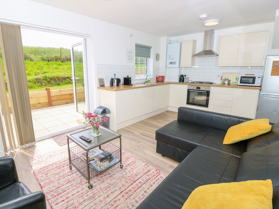A kitchen and living area with black leather sofa yellow cushions a small table with flowers and books and open doors leading outside at 6 Yarmouth Cottages Norton Green near Freshwater
