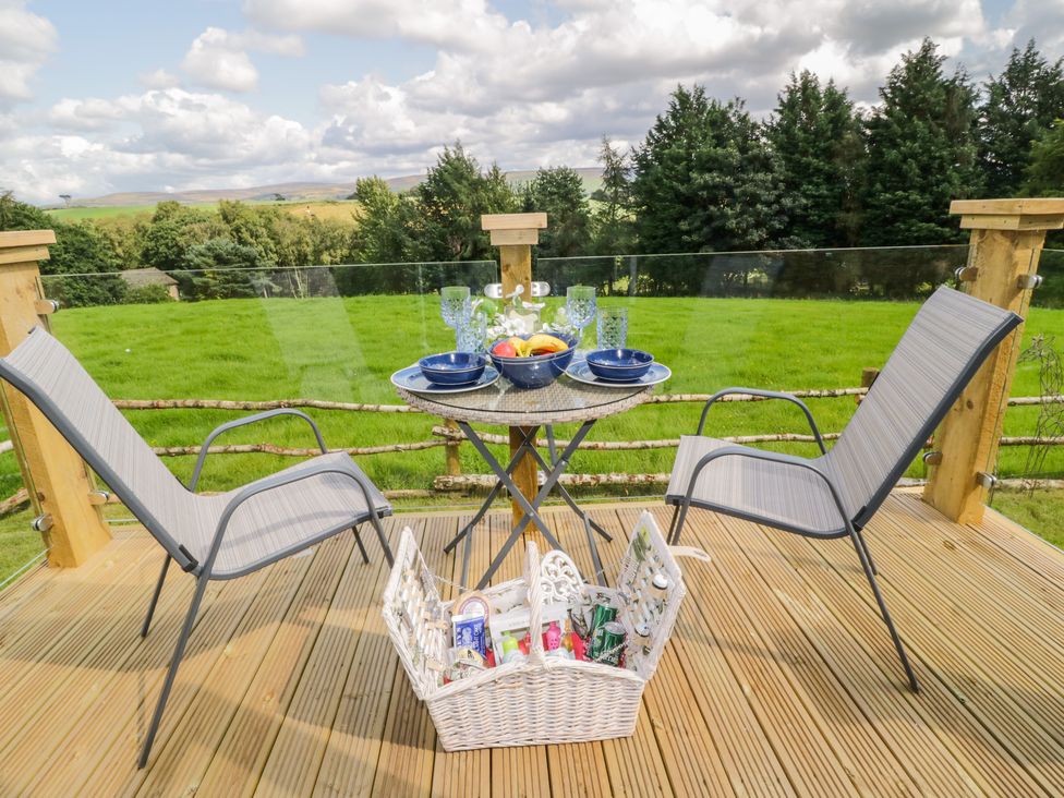 A wooden deck with two chairs a round table set with bowls glasses and fruit with a picnic basket below at High Rigg Shepherd's Retreat in Brampton Cumbria