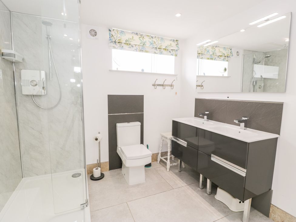 A bathroom with a glass shower enclosure a toilet a double sink vanity large mirror and floral window blinds at Capel Mawr Basement 19 in Llanrug