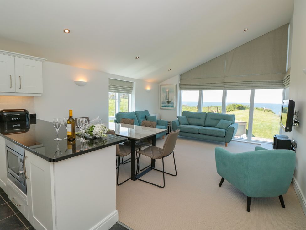 A living room with teal sofas and armchair a dining table and kitchen counter with wine and glasses and large windows overlooking a garden and sea at 7 Ocean's Edge in Thurlestone