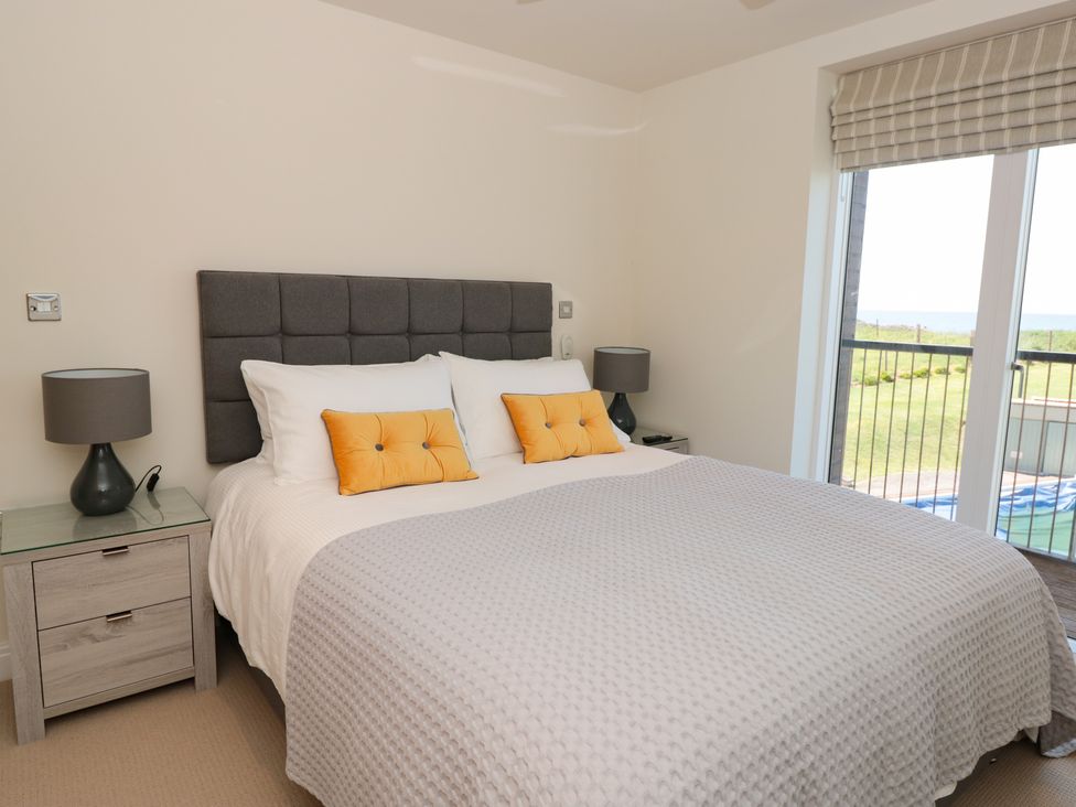 A bedroom with a large bed with gray headboard white bedding and orange pillows two bedside tables with lamps and a sliding door to a balcony at 7 Oceans Edge Thurlestone