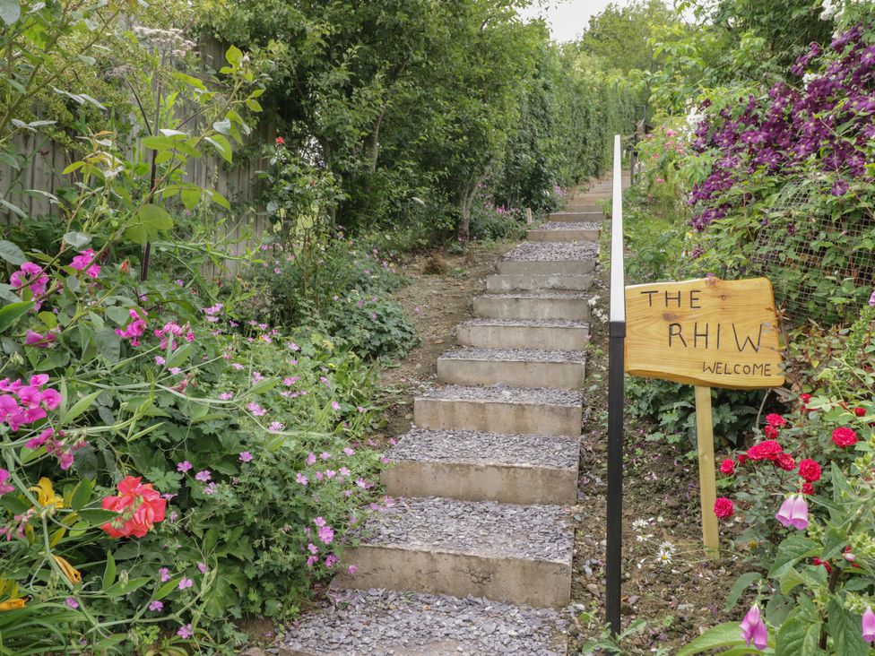 A garden pathway with steps and flowers on both sides and a wooden sign saying The Rhiw welcome at The Rhiw in Llanfair Caereinion