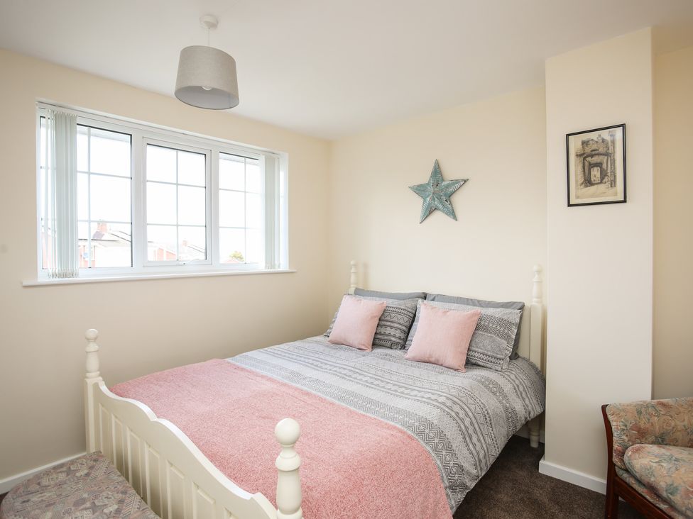 A bedroom with a double bed with pink and gray bedding a window with vertical blinds a wooden framed chair and wall decorations at Yr Hafan in Llysfaen near Llanddulas