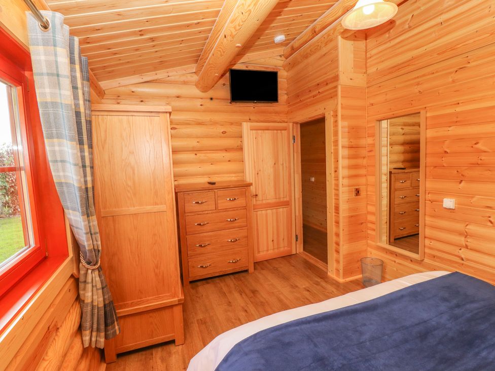 A wooden bedroom with a bed, wardrobe, chest of drawers, wall-mounted tv, mirror, and window with curtains at Maple Lodge in Greetham Rutland