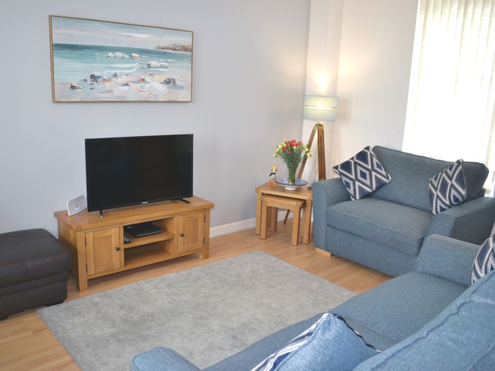 A living room with a television on a wooden stand a sea painting on the wall a floor lamp a bouquet of flowers on nested side tables and blue sofas with patterned cushions at Tammie Norrie Cottage in Banff