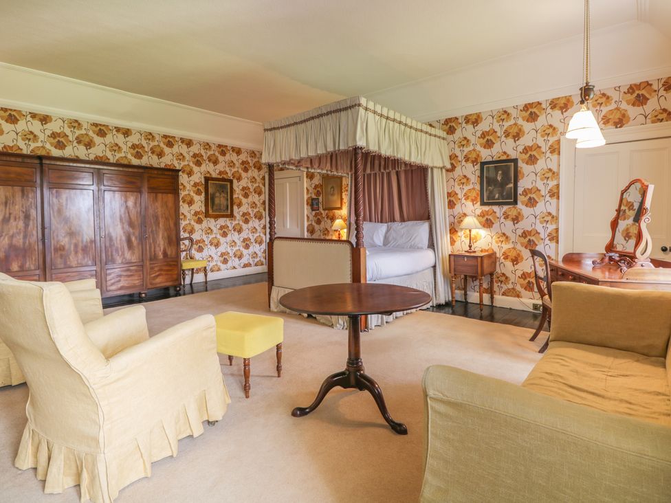 A room with a four poster bed a wooden wardrobe armchairs a small round table and floral wallpaper at The Laird's Wing - Brodie Castle in Forres
