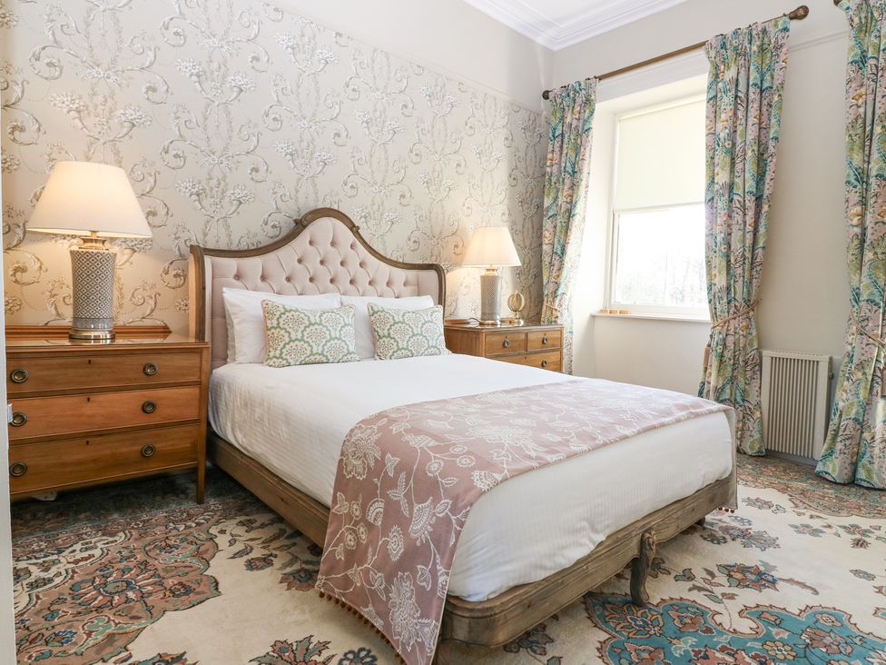 A bedroom with a bed between two wooden nightstands with lamps and a window with floral curtains at North Wing - Pitmedden Gardens in Pitmedden