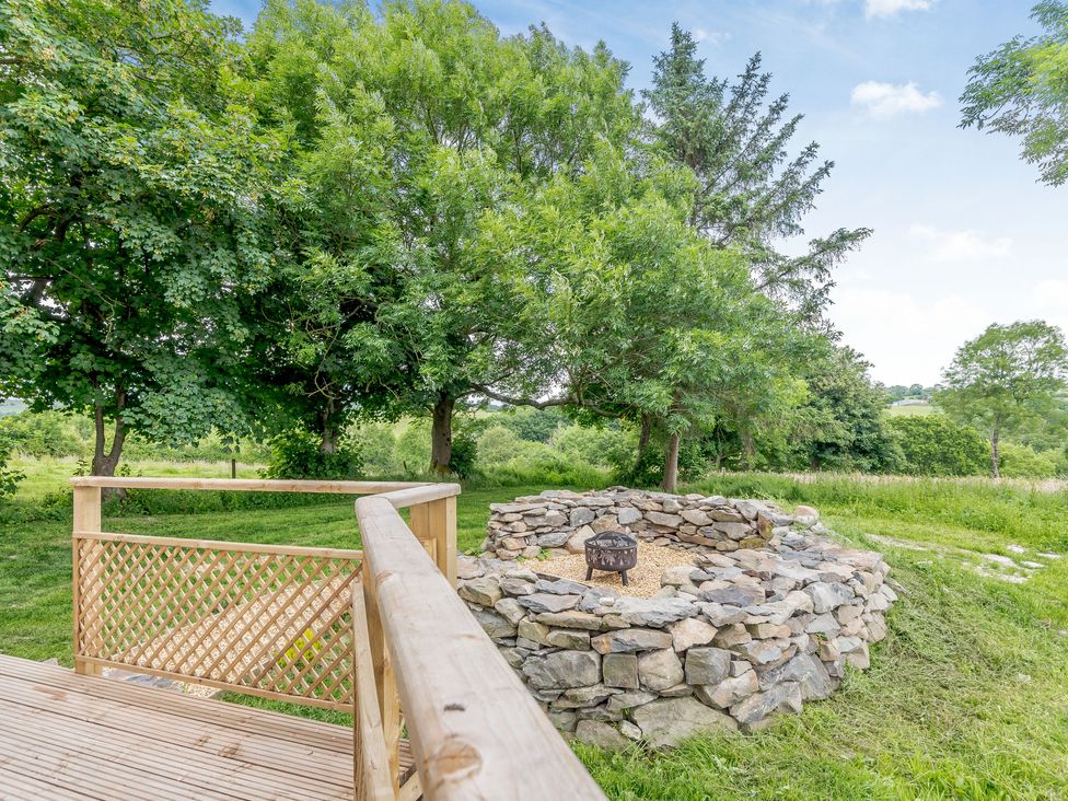 An outdoor stone fire pit surrounded by grass and trees next to a wooden deck with railings at Stag Lodge Pod in Llanfair Caereinion