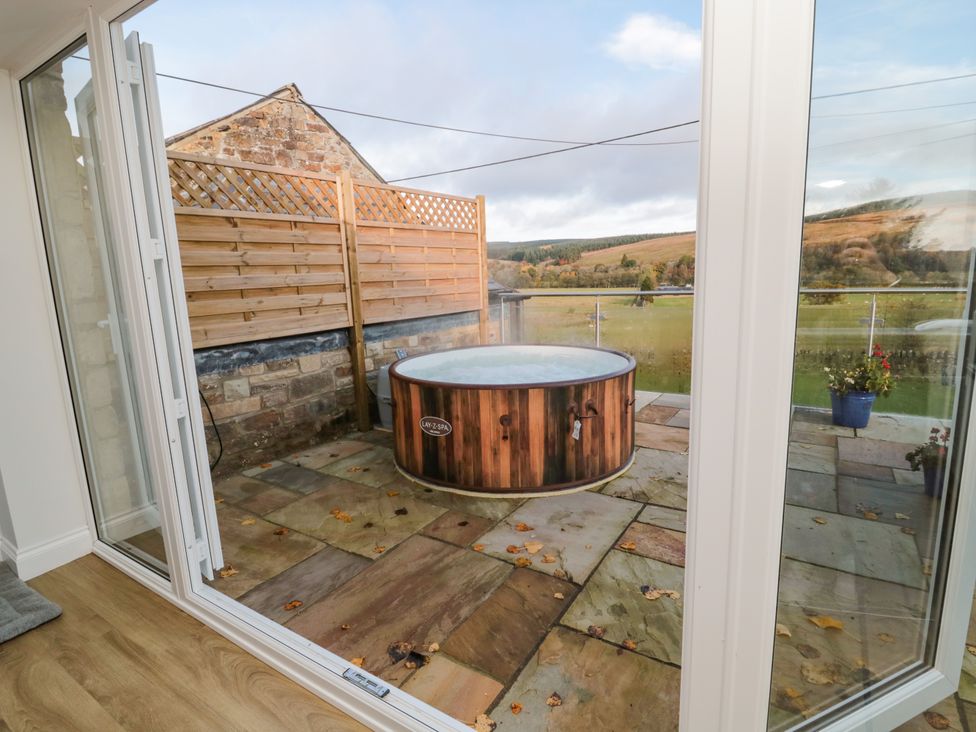 A round wooden hot tub on a stone patio outside a glass door with a wooden fence and countryside in the background at Fox View Cottage in Falstone