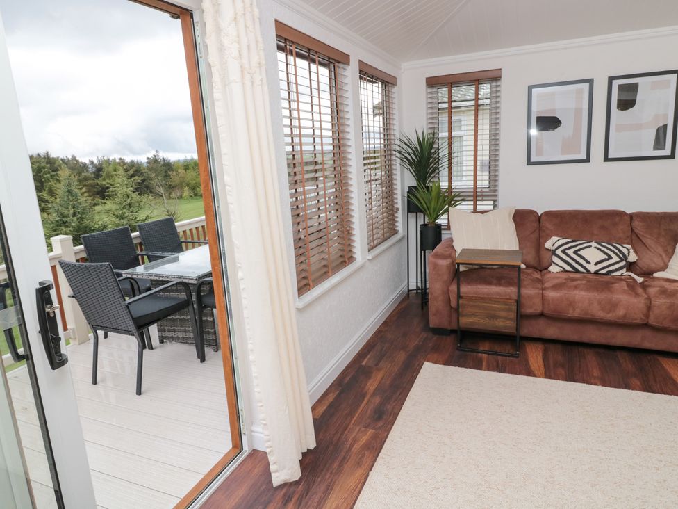 A living room with a brown sofa and pillows next to windows and a sliding door leading to a deck with outdoor chairs and table at Woodland View Lodge in Swarland near Felton