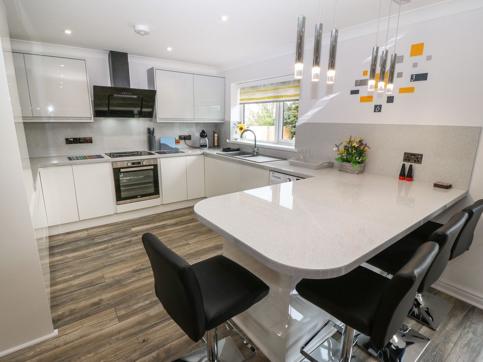 A modern kitchen with white cabinets an oven a sink a large countertop island with black bar stools and pendant lights at Bwthyn Eirlys Snowdrop Cottage in Cemaes Bay