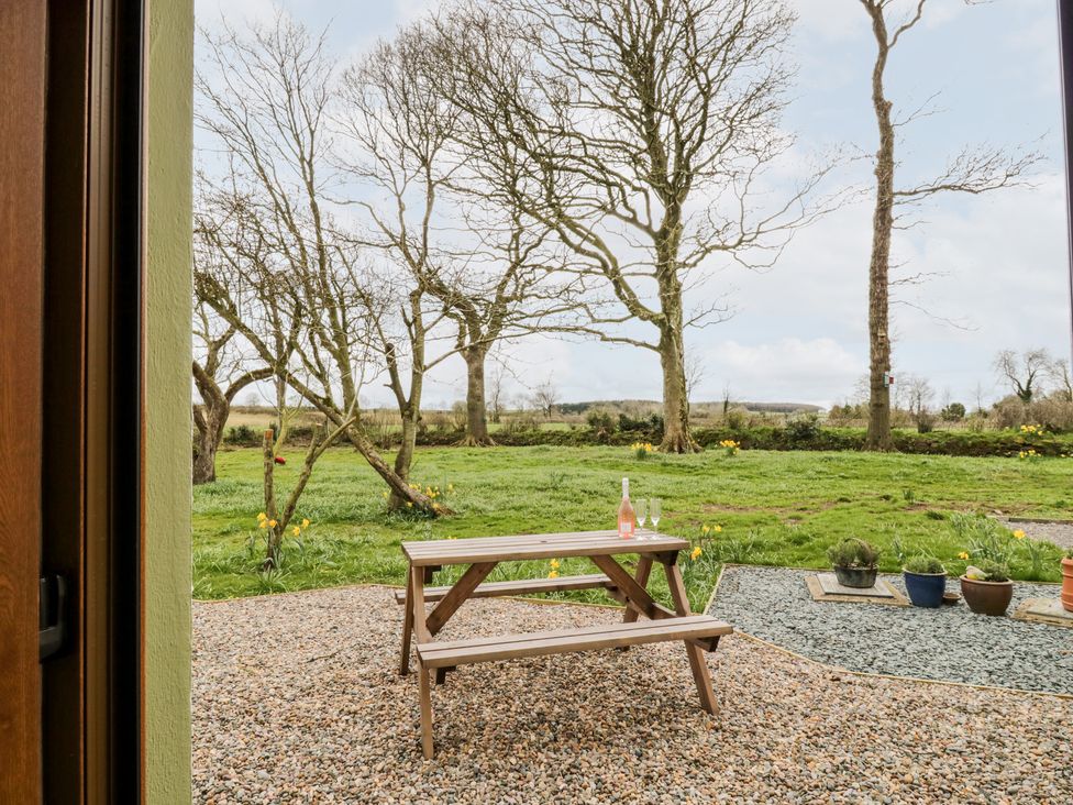 A wooden picnic table with a bottle and two glasses on a gravel patio with trees and grass beyond at Bramblewood Cottage in Crookdake near Bassenthwaite