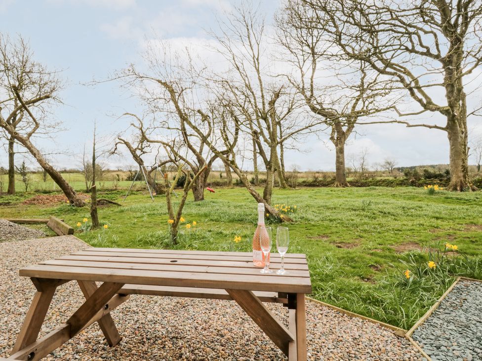A wooden picnic table with a bottle of rose and two glasses on a gravel patio overlooking a grassy garden with trees and a swing at Bramblewood Cottage in Crookdake near Bassenthwaite