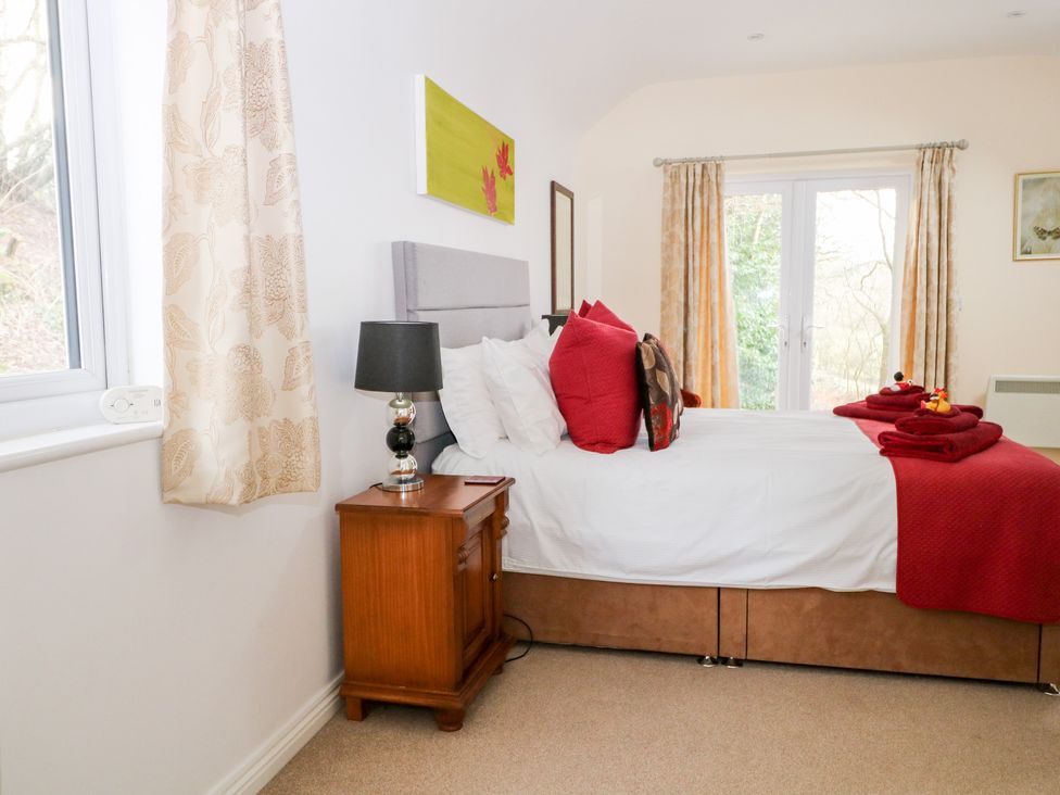 A bedroom with a bed with red pillows and blankets a wooden bedside table with a lamp and windows with curtains at Heath Retreat in Buxted