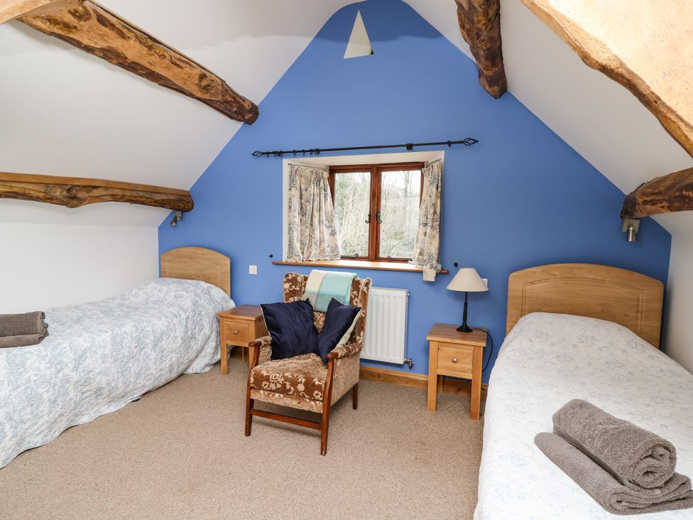 A bedroom with two single beds a window in the center two wooden nightstands a chair with pillows and exposed wooden beams at Maes Y Berllan Barn in Gilwern